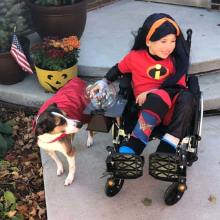 small girl in wheelchair wearing an Incredibles costume with her dog next to her in a cape