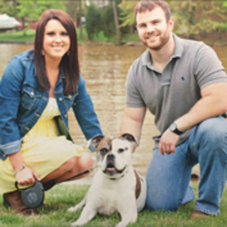 a man and a women sitting with a multi colored dog in front of lake